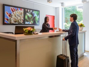 Un comptoir d’accueil avec un écran affichant des images de fleurs, une plante en pot et une fenêtre donnant sur un paysage verdoyant.