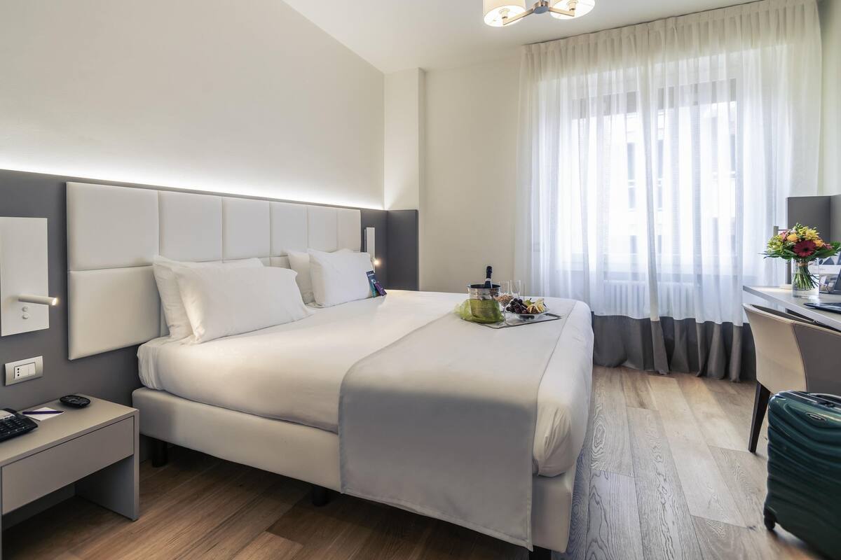 A modern hotel room with a large bed, bedside table, desk, and a view of the city through sheer curtains.