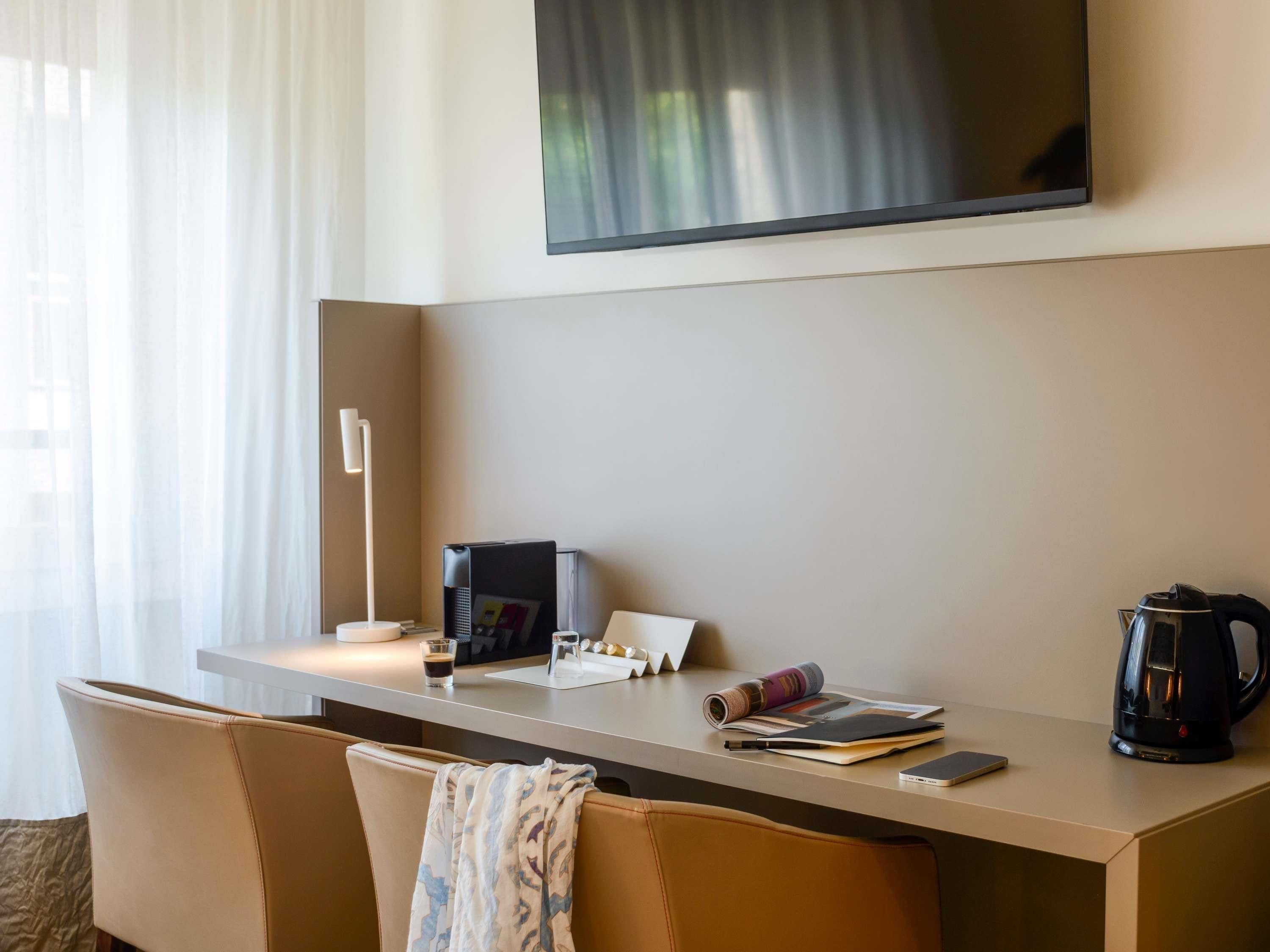 Une chambre d’hôtel équipée d’un bureau, d’une chaise, d’un téléviseur fixé au mur, d’une machine à café et d’une fenêtre avec des rideaux.