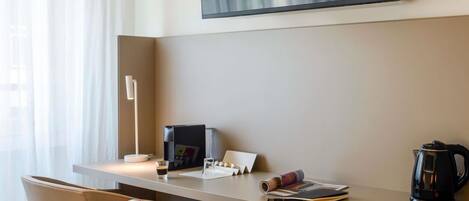Une chambre d’hôtel équipée d’un bureau, d’une chaise, d’un téléviseur fixé au mur, d’une machine à café et d’une fenêtre avec des rideaux.