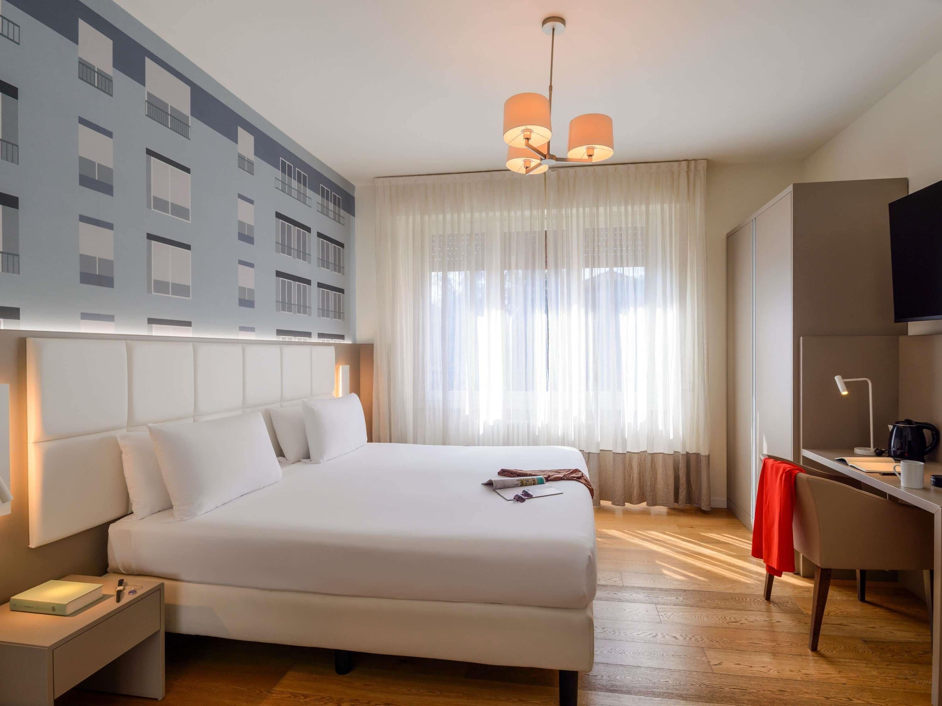 Una habitación de hotel con una cama grande, un escritorio, un televisor y una ventana con cortinas translúcidas.