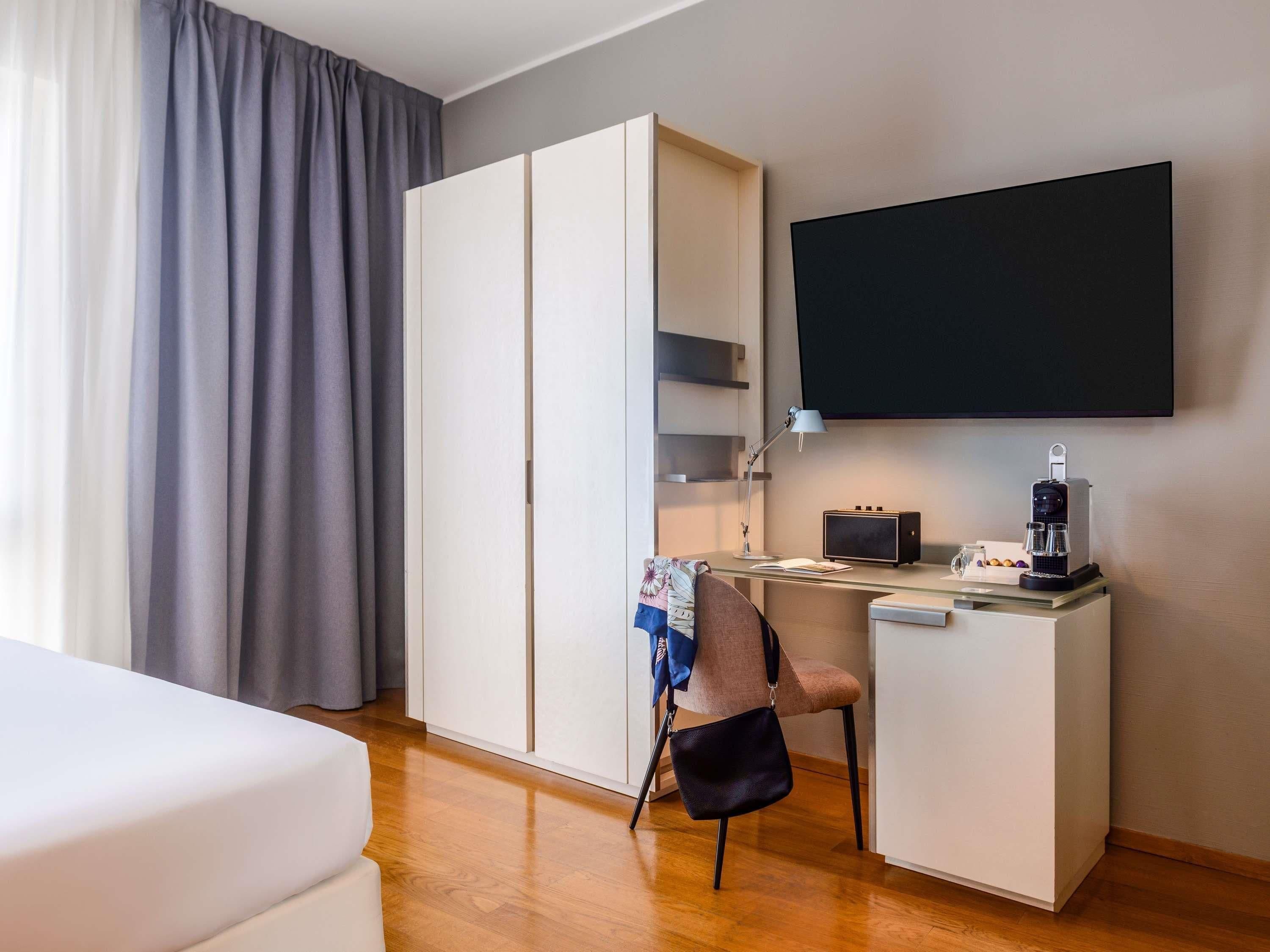 A hotel room with a bed, a desk with a chair, a wardrobe, a television, and a coffee machine.
