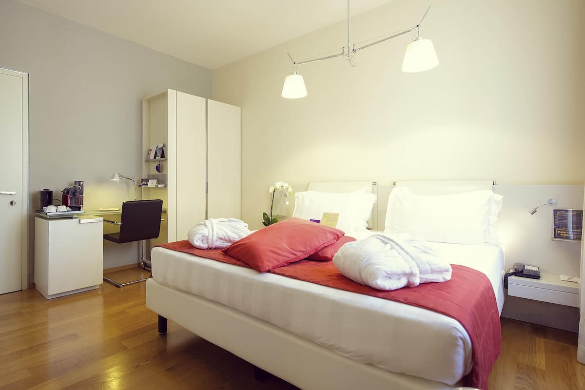 A hotel room with a bed, bedside table, desk, wardrobe, and a desk lamp.