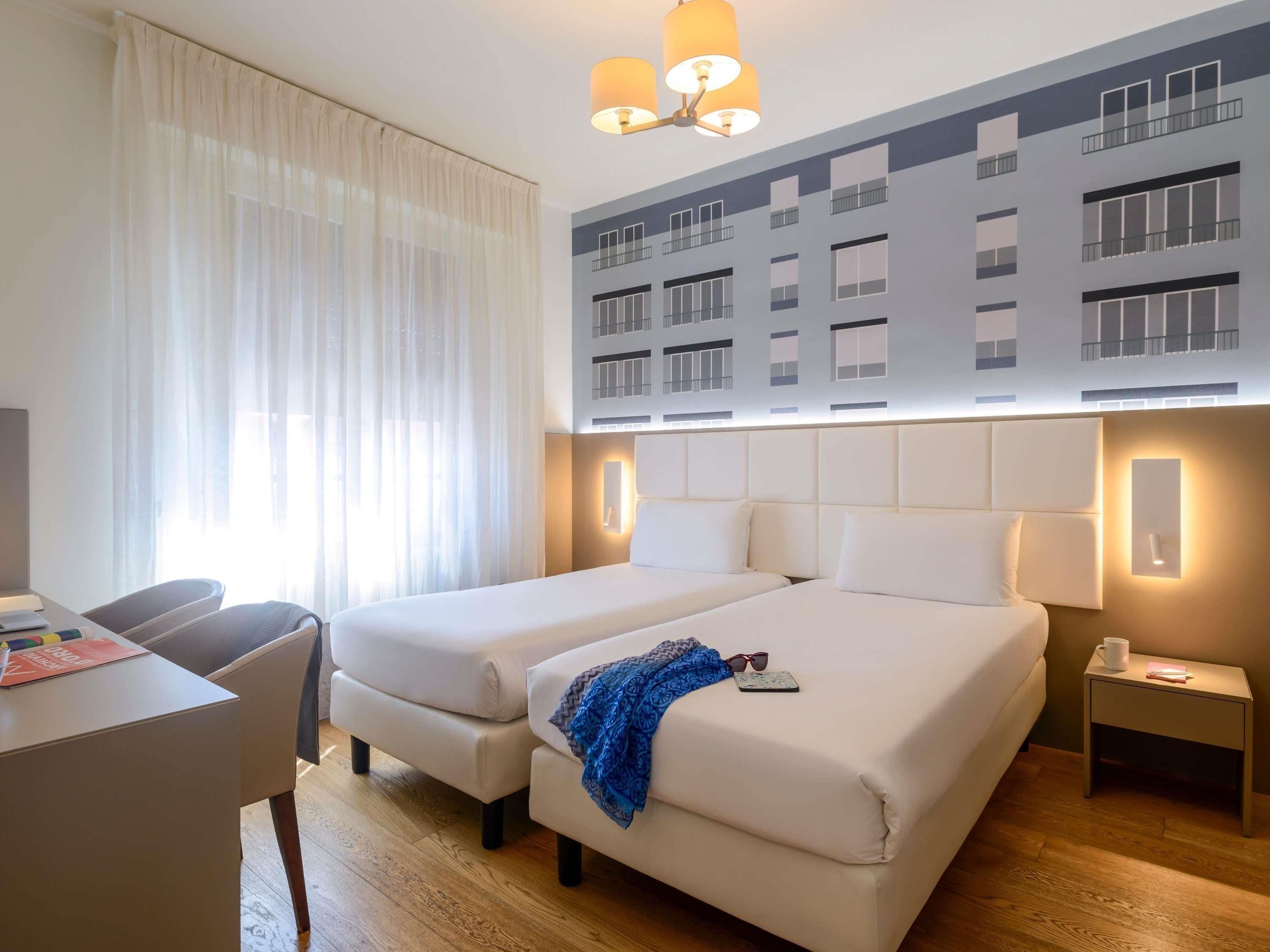 A hotel room with two beds, a desk, a chair, a nightstand, and a large window with curtains.