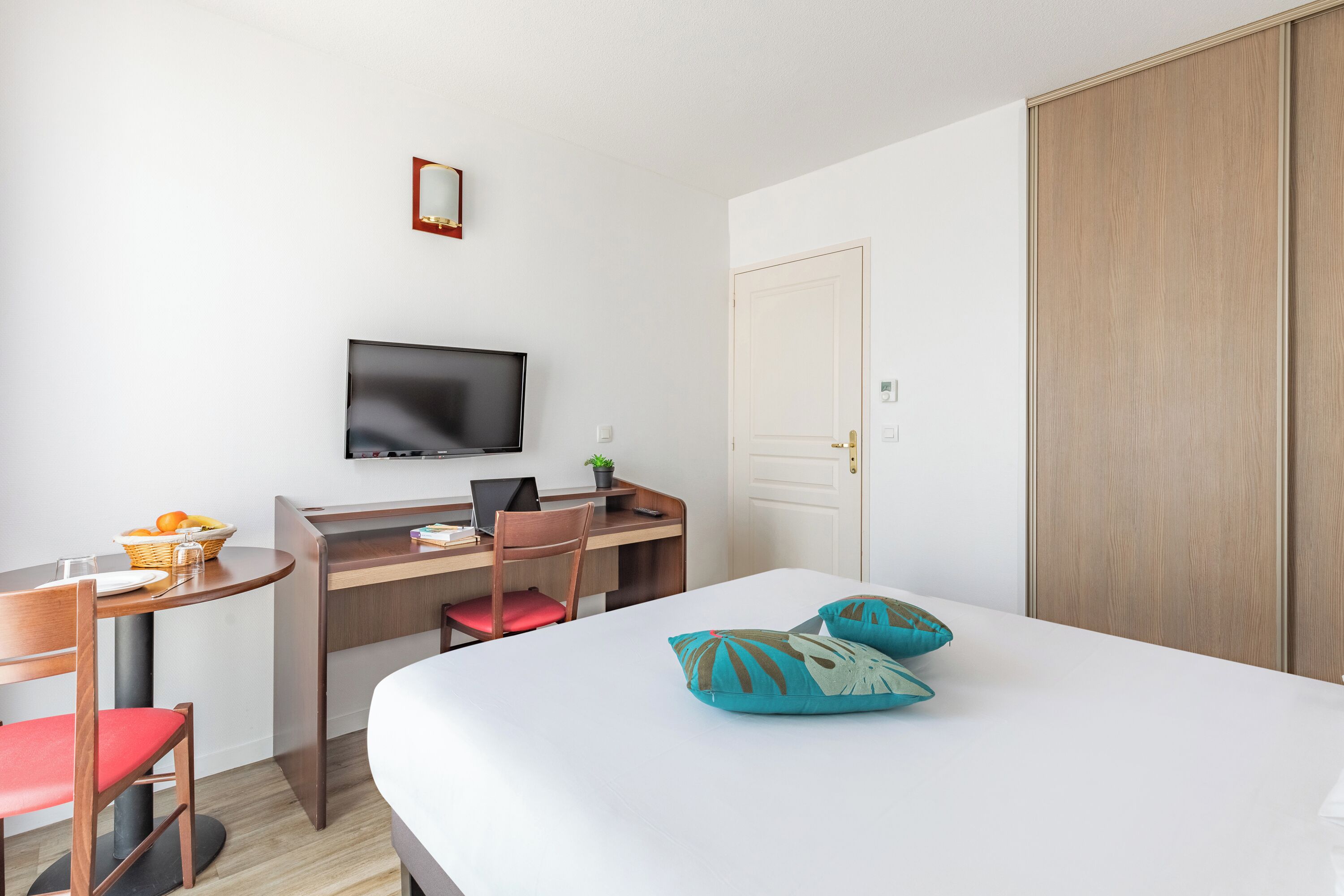 A hotel room with a bed, a desk with a computer, a chair, a television, and a wardrobe.
