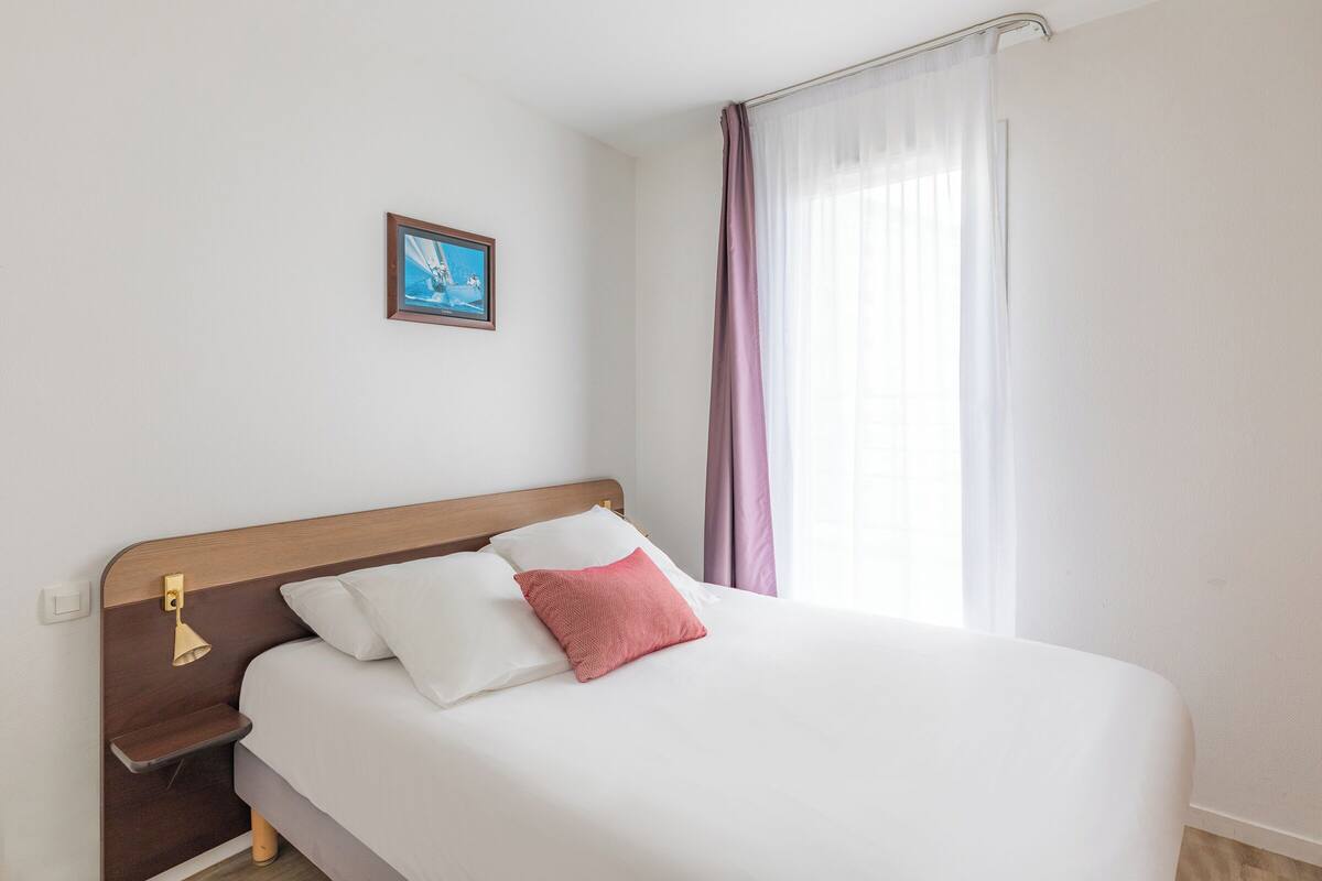 Een keurig opgemaakt bed met witte en roze kussens, een klein nachtkastje met een lamp, en een ingelijst foto aan de muur.