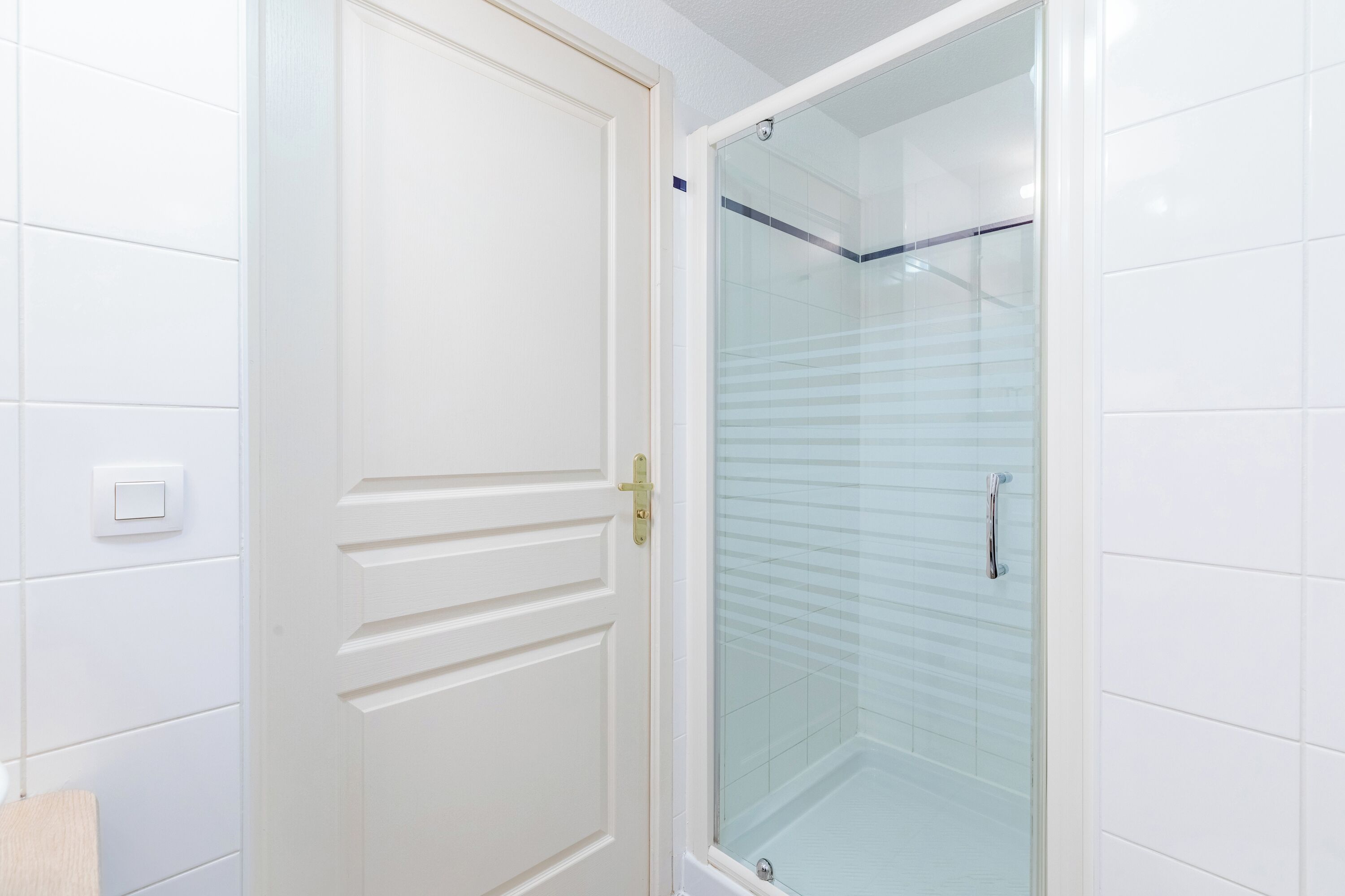 A bathroom with white tiled walls, a white door with a brass handle, and a glass shower enclosure.