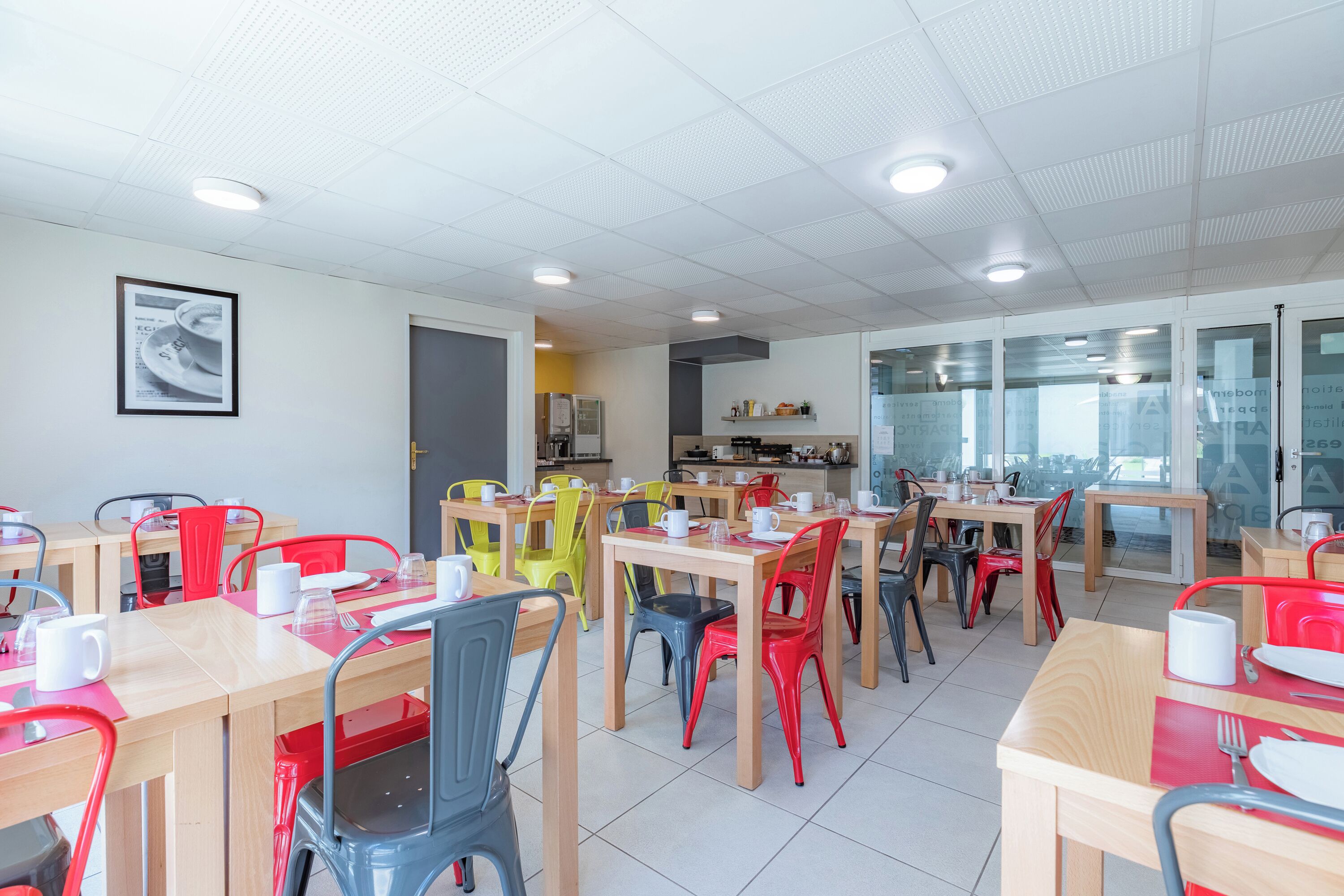 A modern dining area with multiple tables and chairs, a kitchen in the background, and a framed picture on the wall.