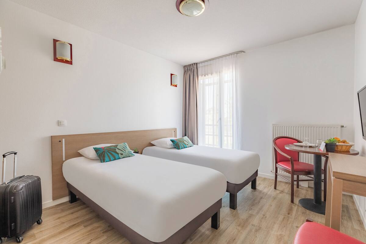 Hotelkamer met twee bedden, een bureau, een stoel, een klein tafeltje, een raam met gordijnen en een radiator.