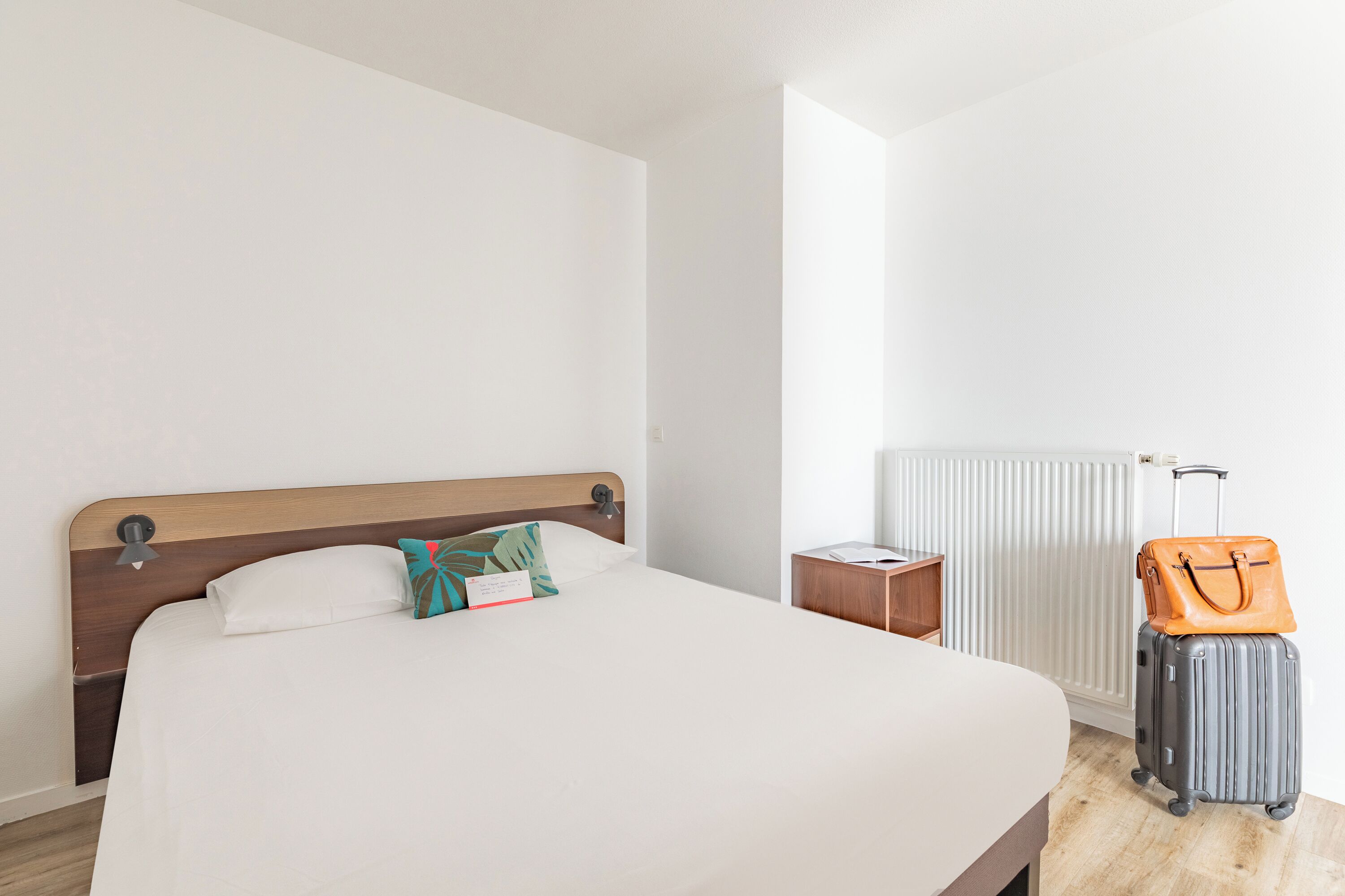 A neatly made bed with a wooden headboard, a small wooden nightstand, a radiator, and a suitcase with a handbag on top.