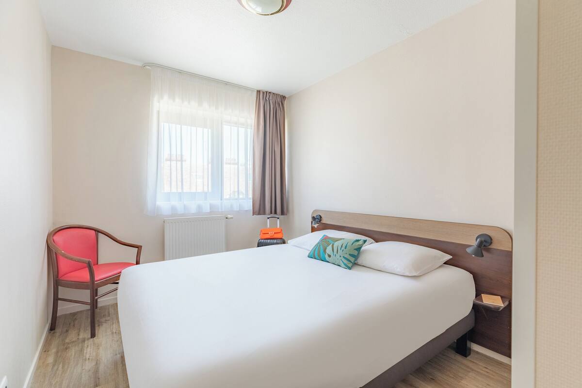 Een hotelkamer met een groot bed, een rode stoel, een nachtkastje met een lamp en een raam met gordijnen.