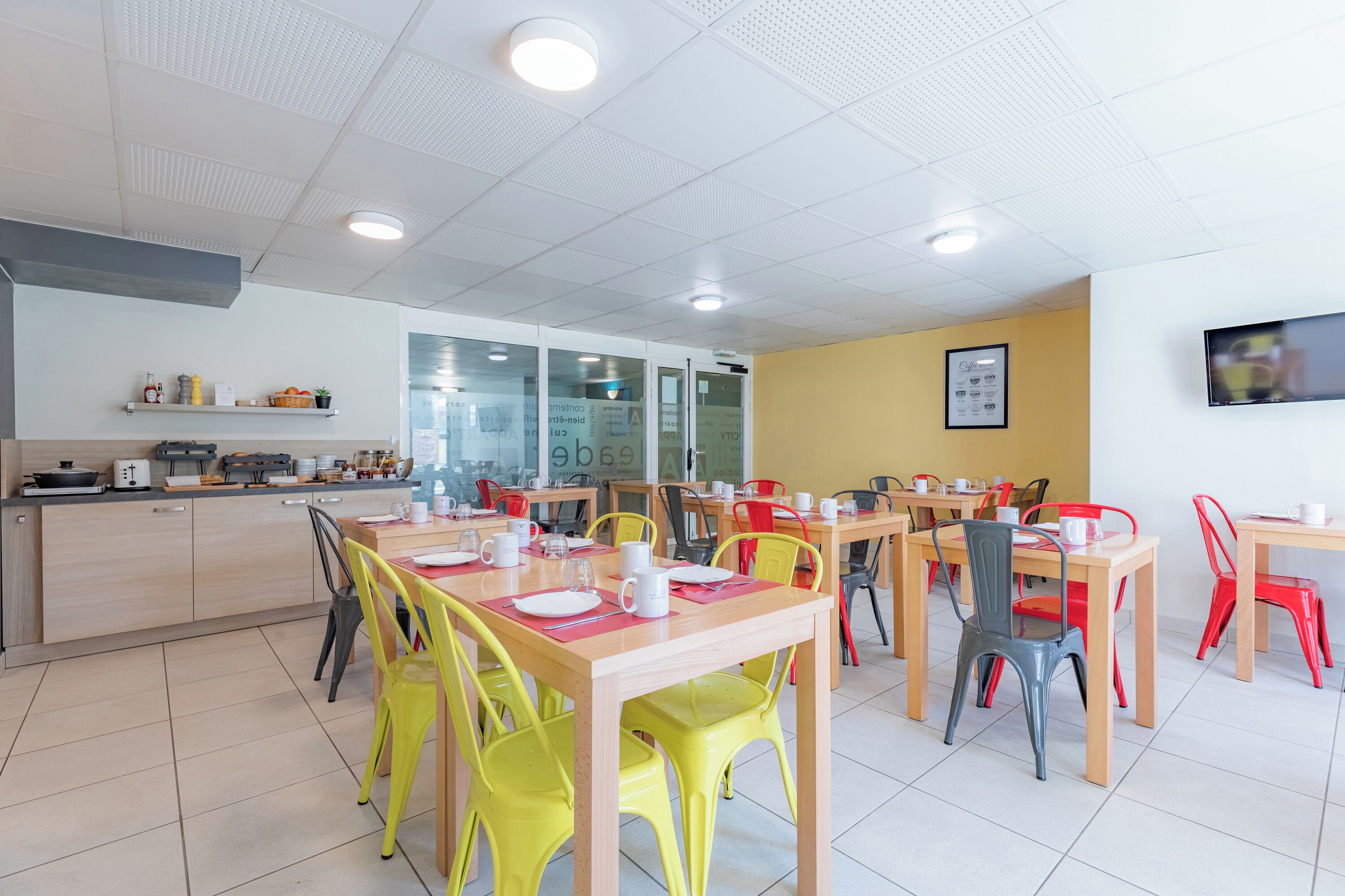 A modern dining area with multiple tables and chairs, a kitchen area with appliances, and a TV mounted on the wall.