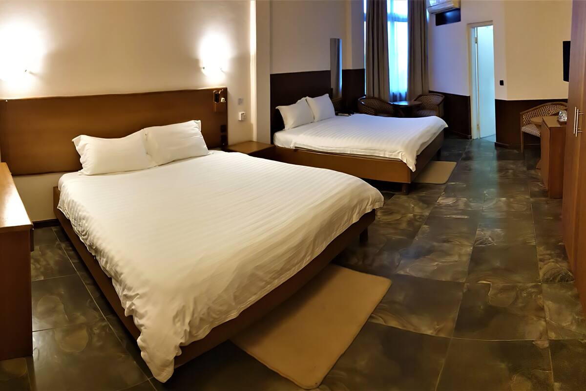 A hotel room with two beds, a wooden nightstand, a small table, and a chair. The room has a tiled floor and a door leading to another room.