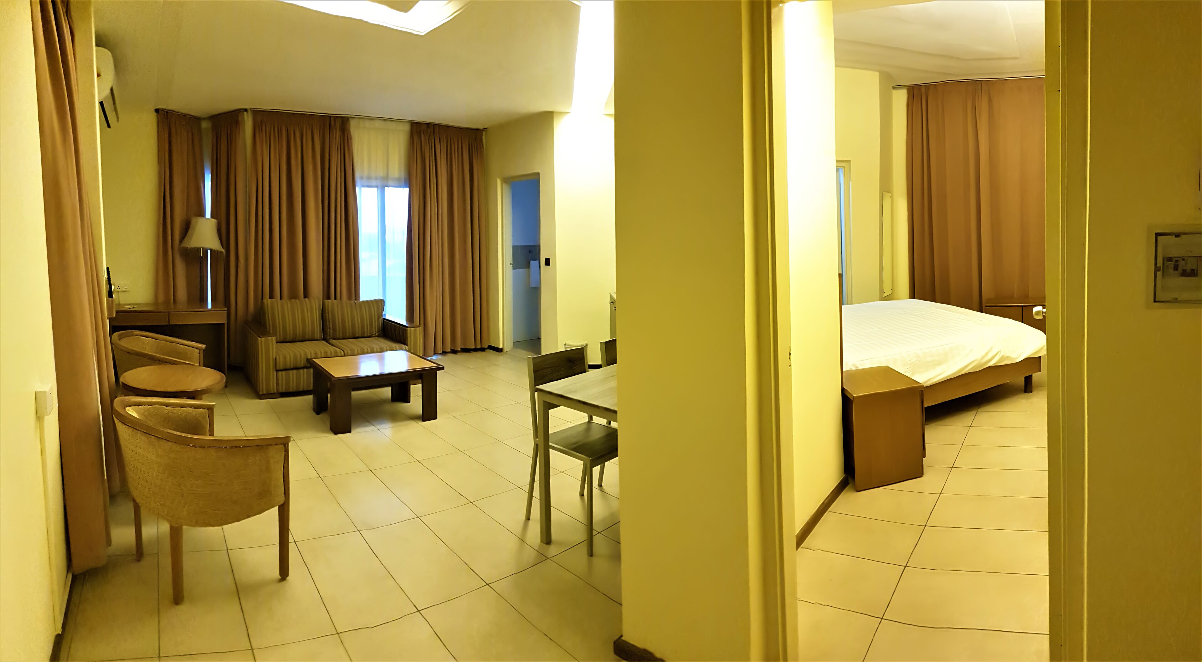 A hotel room with a bed, a sofa, a small table, and two chairs. There is a window with curtains, and the room has a tiled floor.