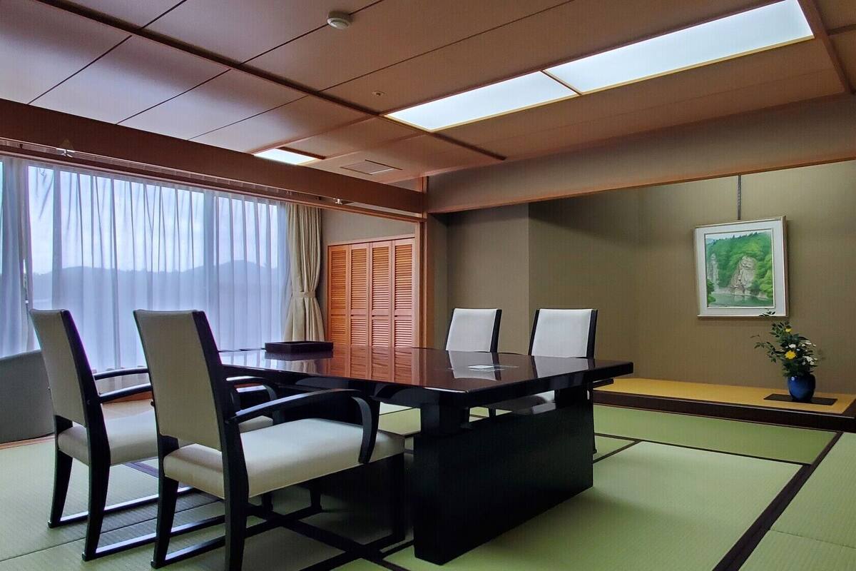 A conference room with a large table, chairs, a tatami mat floor, a window with curtains, a door, and a painting on the wall.