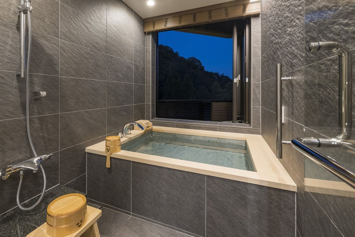 A modern bathroom with a bathtub, shower, and a window overlooking a mountainous area.