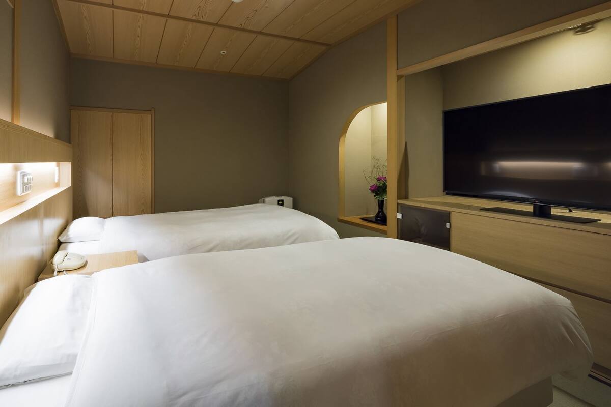 A hotel room with two beds, a TV, a bedside table with a phone, a mirror, and a wall-mounted heater.