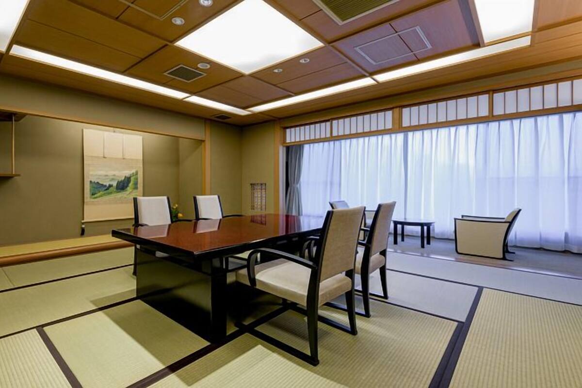 A conference room with a wooden table, chairs, tatami mats, a painting, and a large window with curtains.