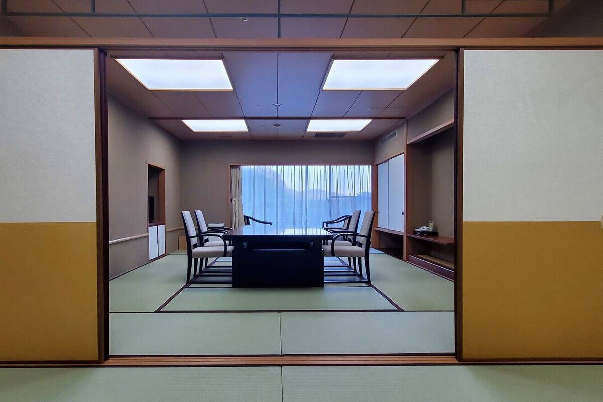 A traditional Japanese room with tatami flooring, a low table, and chairs, featuring a large window with a view of the sea.