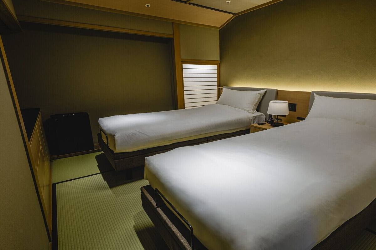 A hotel room with two beds, a bedside table with a lamp, a small TV, and a sliding door.