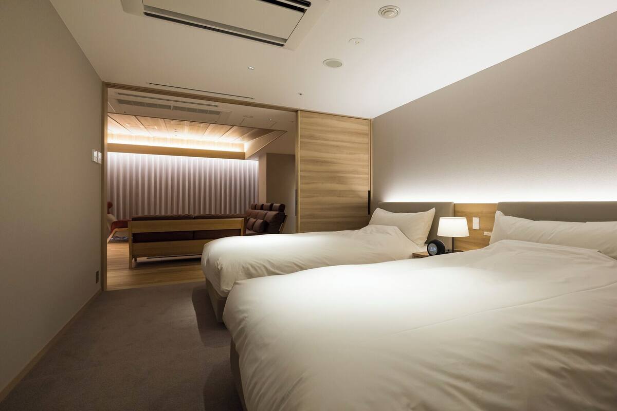 A hotel room with two beds, a bedside table with a lamp, a wooden headboard, and a sliding door leading to a seating area.