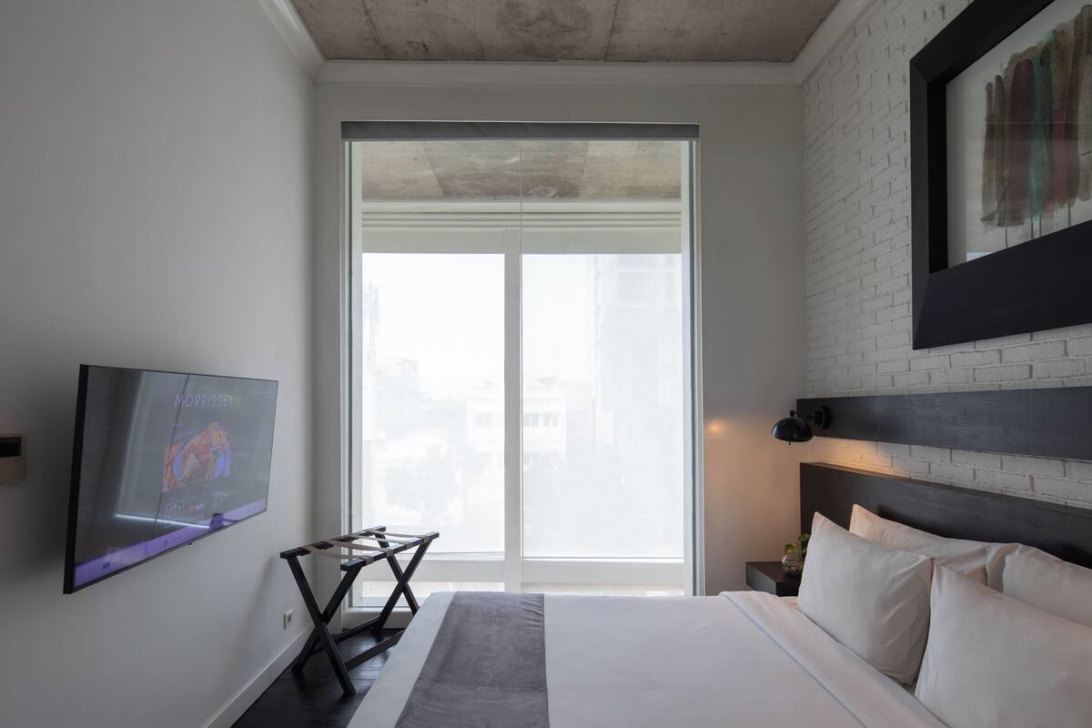 A modern hotel room with a large bed, a flat-screen TV mounted on the wall, a window with a view of buildings, and a brick wall.