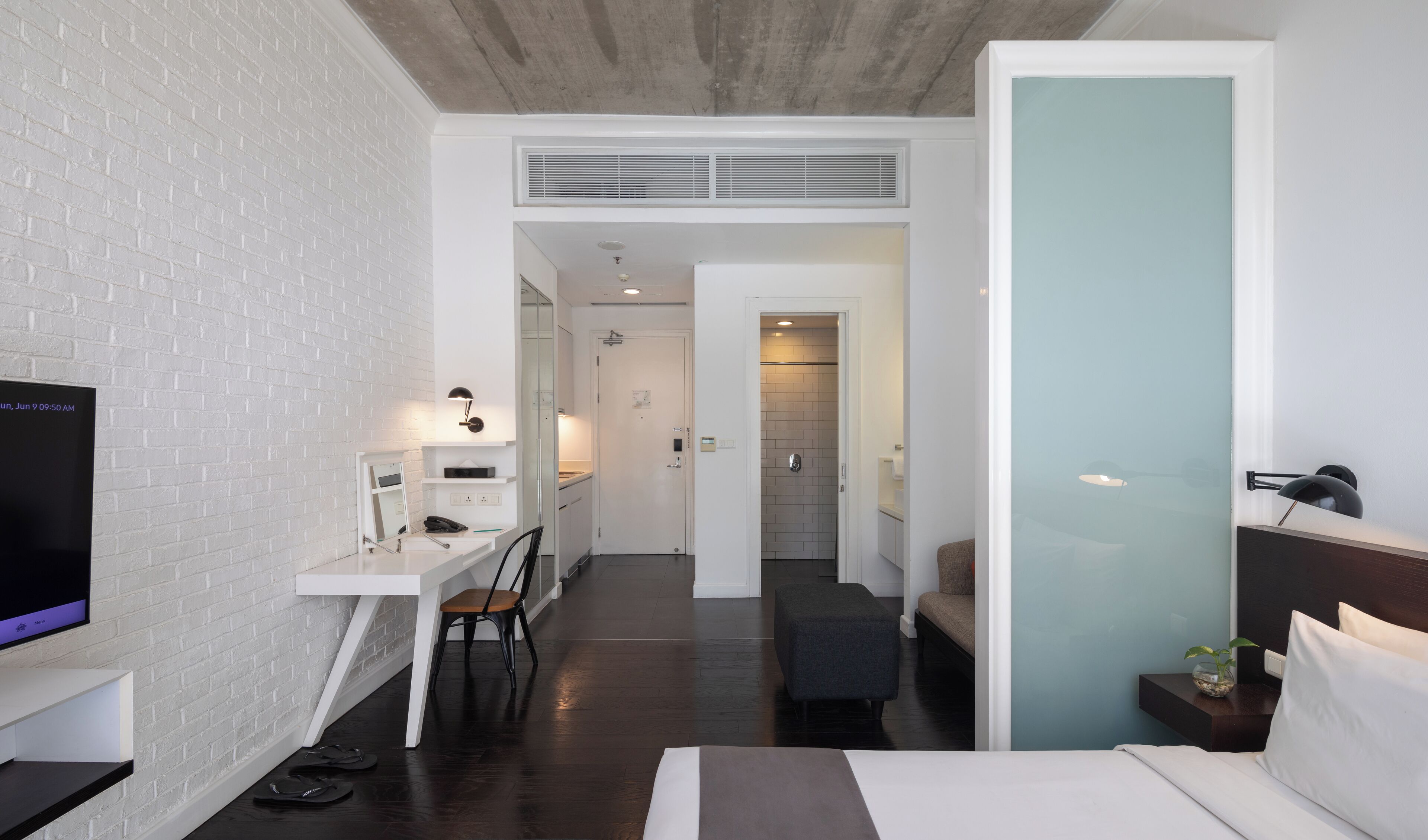 Une chambre d’hôtel moderne avec un mur en briques blanches, un téléviseur à écran plat, un bureau avec une chaise, un lit et une petite table avec une lampe.