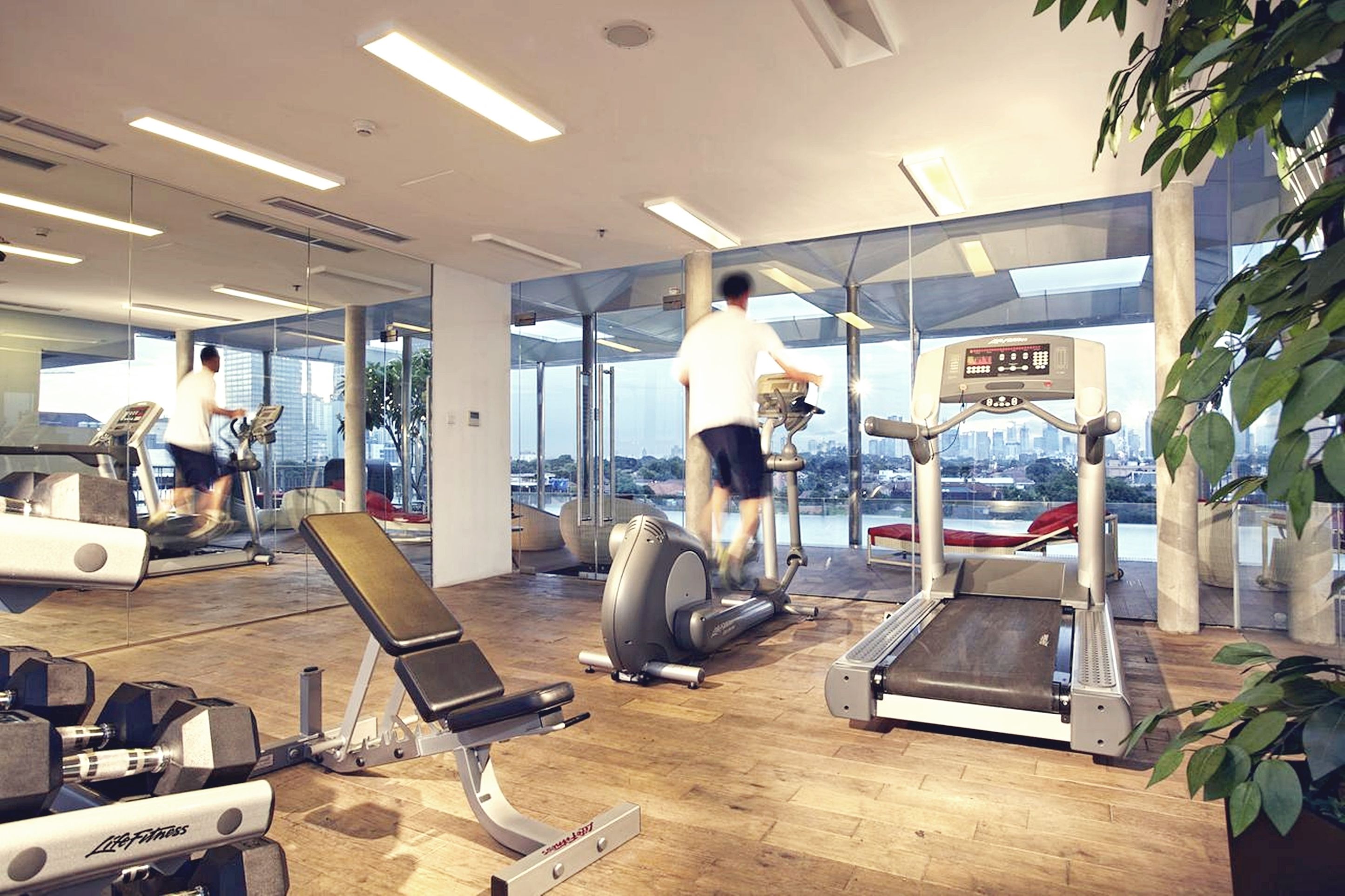 Un gimnasio con diversos equipos de ejercicio, ventanas amplias y vistas de la ciudad.