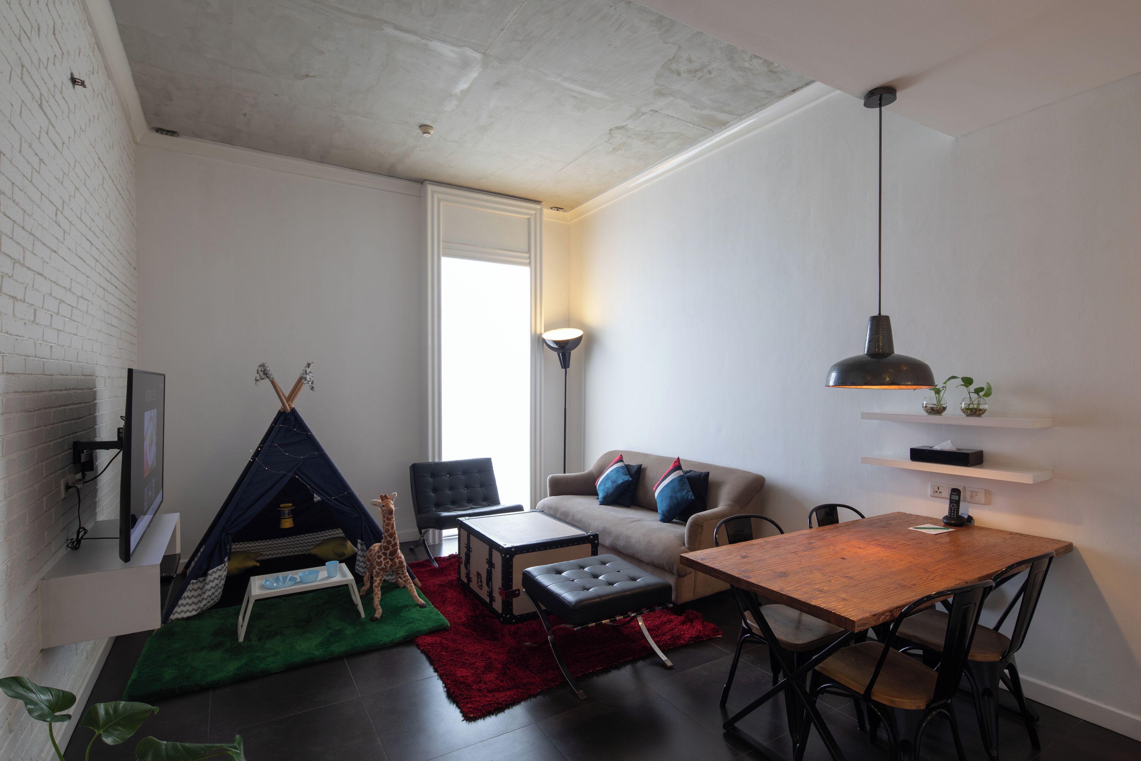 A modern living room with a sofa, a dining table, a red rug, a black leather ottoman, a black chair, a white wall, a white ceiling, a black floor lamp, a white ceiling light, a black TV, and a white door.
