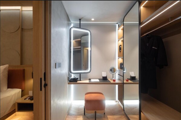 A modern hotel room with a wooden desk, a mirror, a vanity with a round mirror, and a closet with hanging clothes. - Morrissey Hotel (Jakarta)