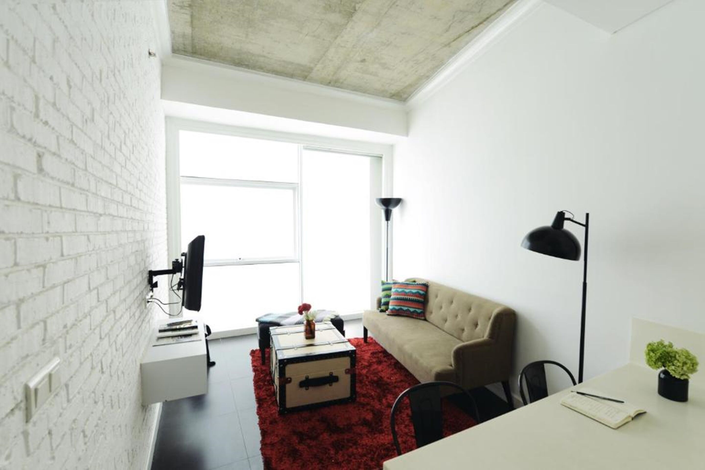 Una sala de estar moderna con una pared de ladrillo blanco, un ventanal grande, un sofá color beige, un tapete rojo y un escritorio con una planta.