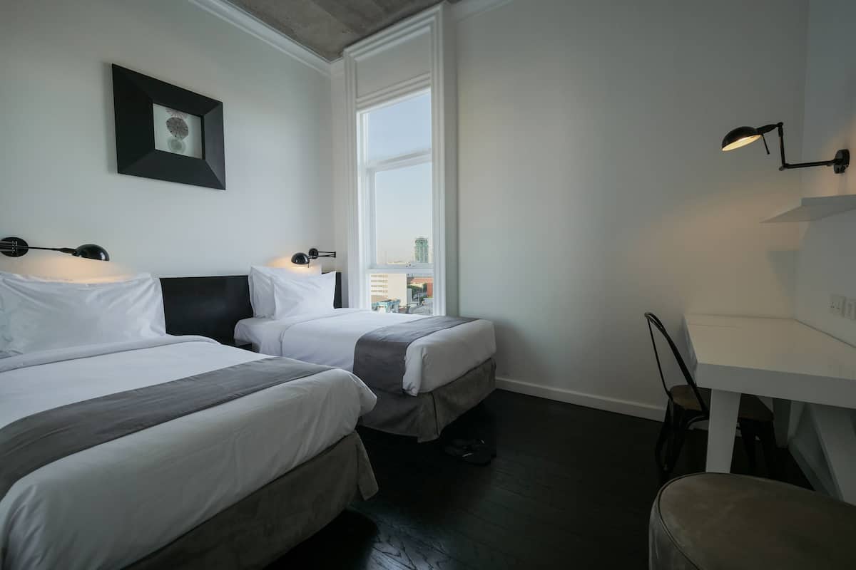 A hotel room with two beds, a desk, a chair, a window, and a picture on the wall.