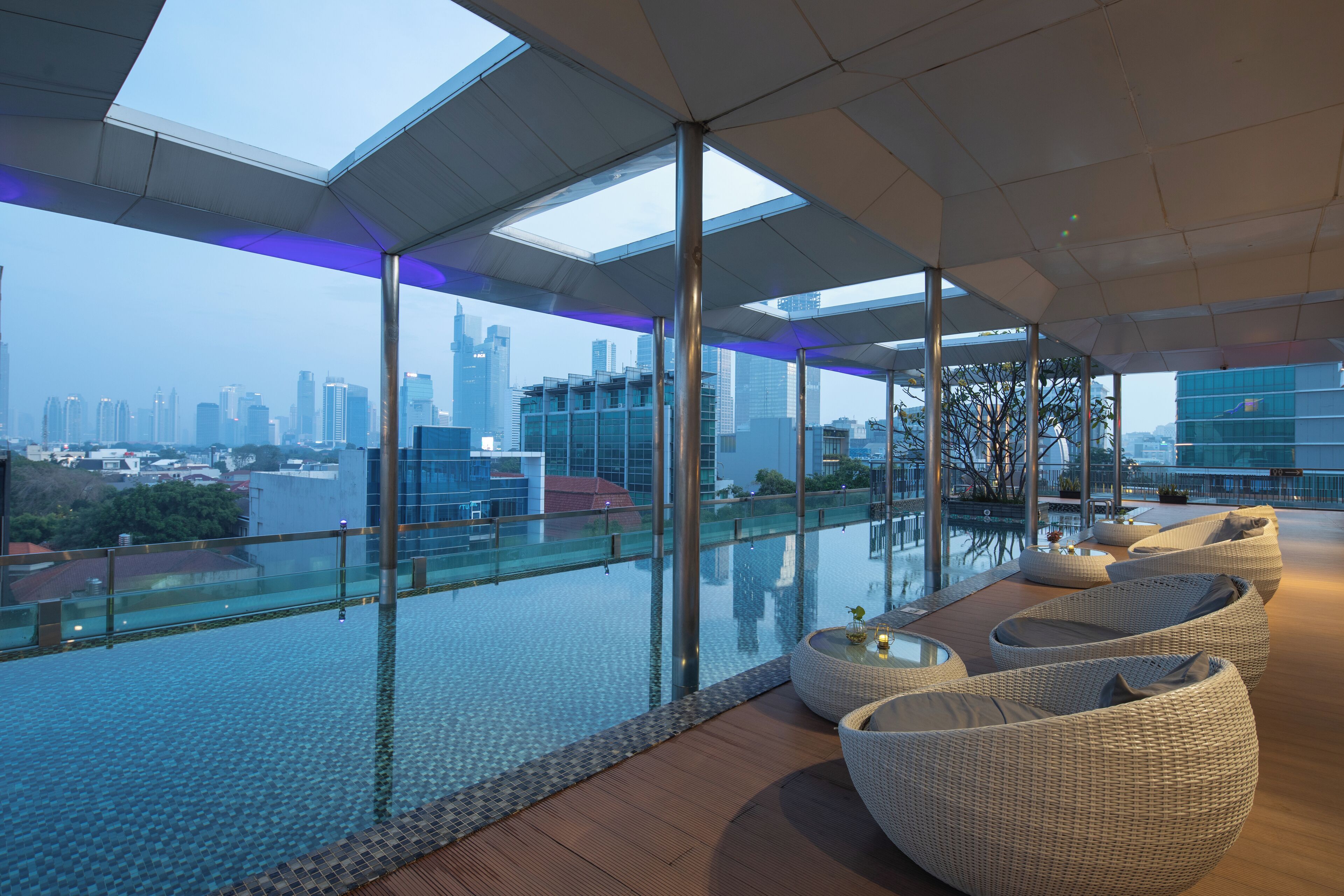 A rooftop infinity pool with glass railings, wicker lounge chairs, and a city skyline view.