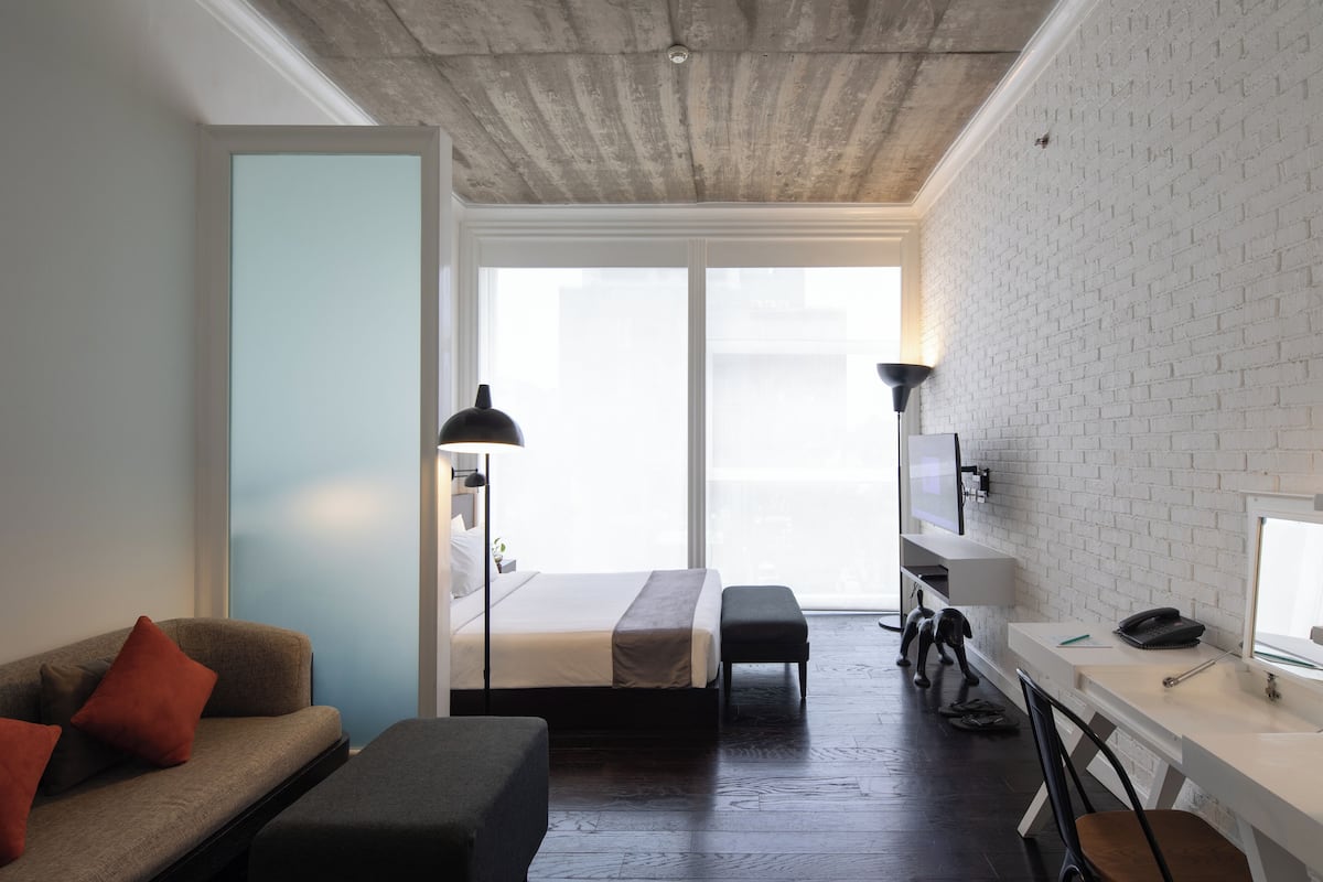 A modern hotel room with a bed, a desk, a chair, a sofa, and a large window.