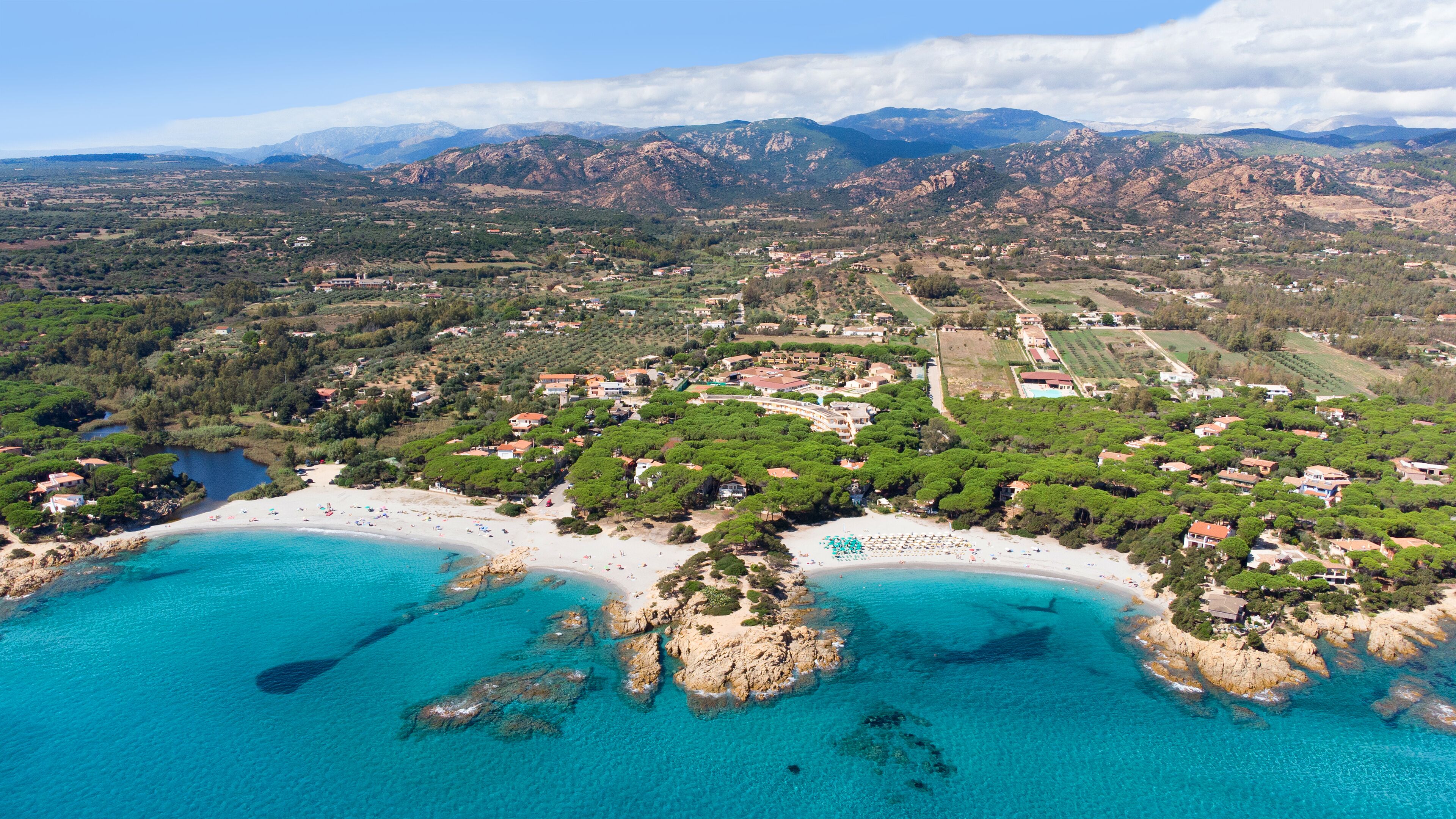 An aerial view of a coastal town with a beach, clear turquoise waters, and mountainous terrain in the background.