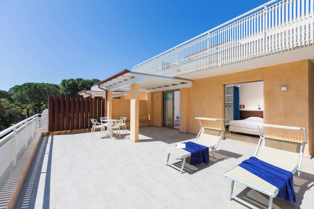 A modern outdoor balcony with lounge chairs and a table, a bedroom with a bed and a bathroom, and a clear blue sky.