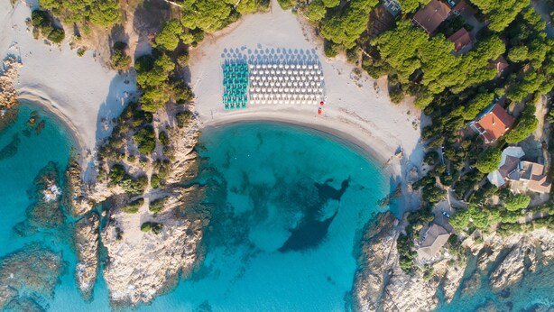 An aerial view of a beach with a row of umbrellas and lounge chairs, rocky coastline, and nearby houses. - Sentido Tirreno Resort (Orosei)