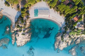 An aerial view of a beach with a row of umbrellas and lounge chairs, rocky coastline, and nearby houses. - Sentido Tirreno Resort (Orosei)