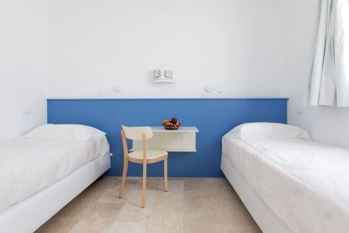 A small, clean room with two single beds, a desk with a chair, and a wall-mounted shelf with a basket of fruit.