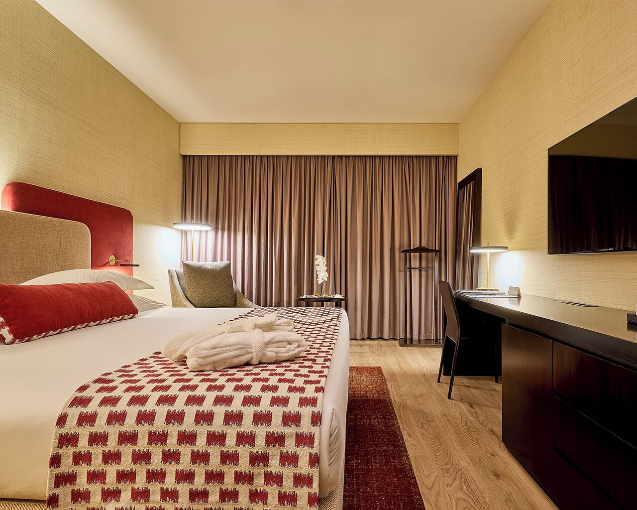 Habitación de hotel con cama, escritorio, silla, televisor y ventana con cortinas.