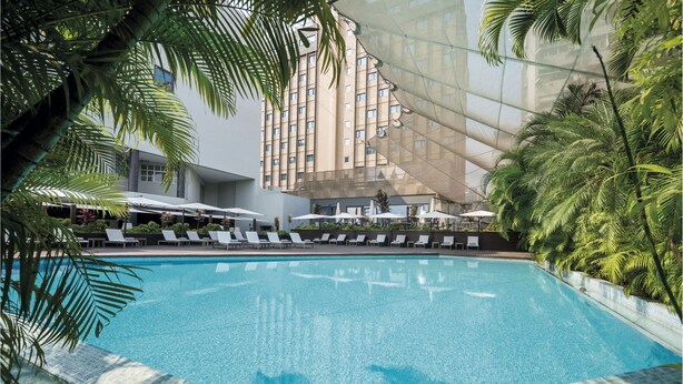 A swimming pool with lounge chairs and umbrellas, surrounded by palm trees and a modern building in the background. - Hotel Trópico (Luanda)