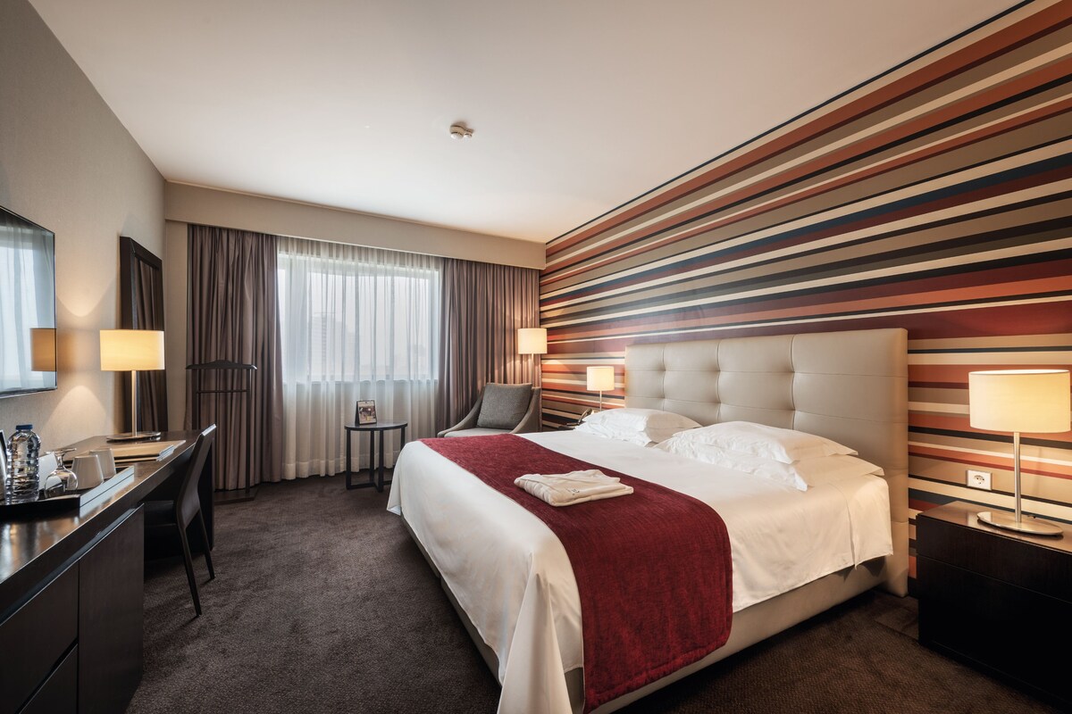 A hotel room with a large bed, a desk, a chair, and a TV.