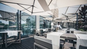 An outdoor dining area with tables and chairs, large umbrellas, and a view of the city through glass walls. - Hotel Trópico (Luanda)