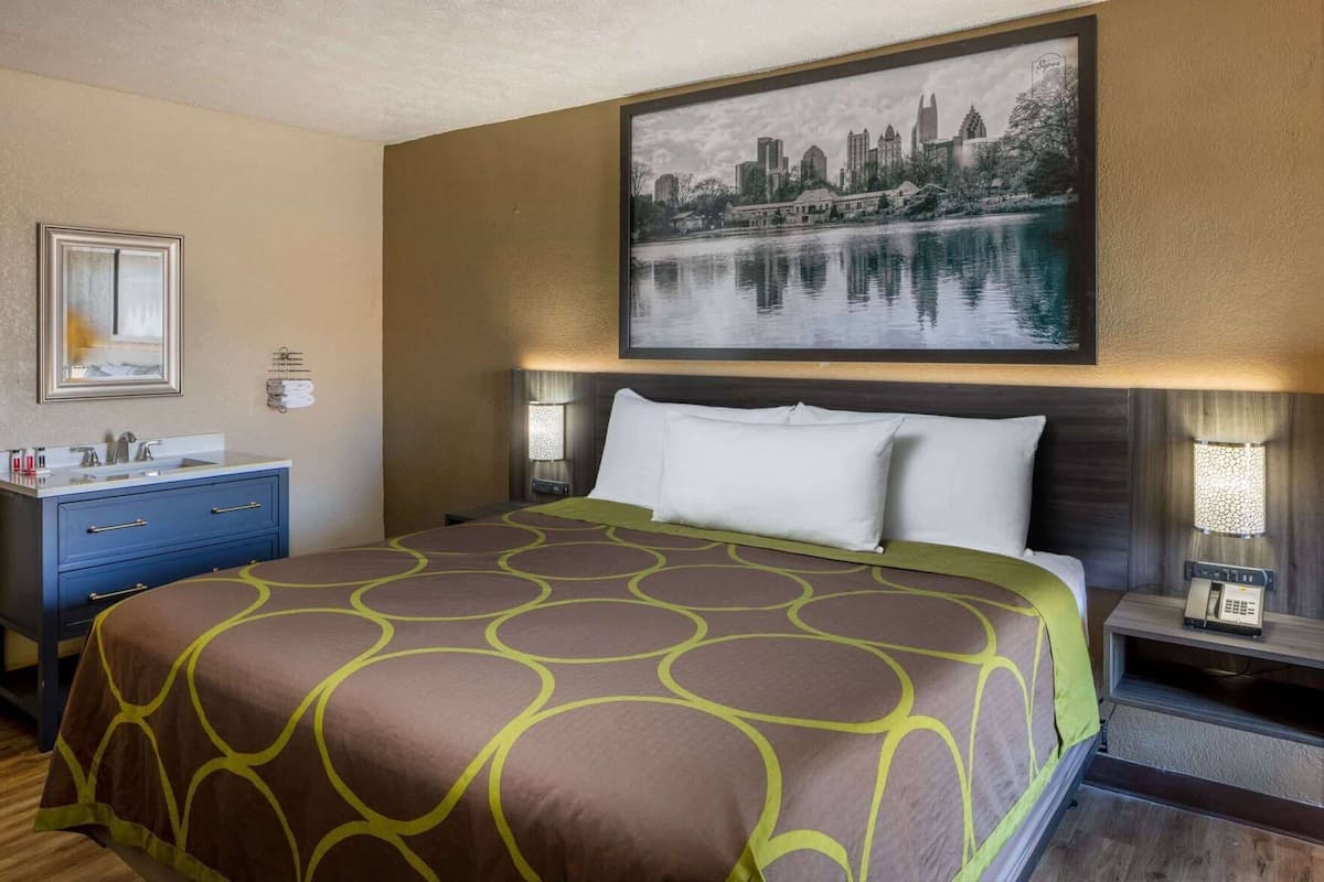 Une chambre d’hôtel avec un lit, une table de chevet, une lampe, un miroir, une commode et un tableau représentant un paysage urbain accroché au mur.