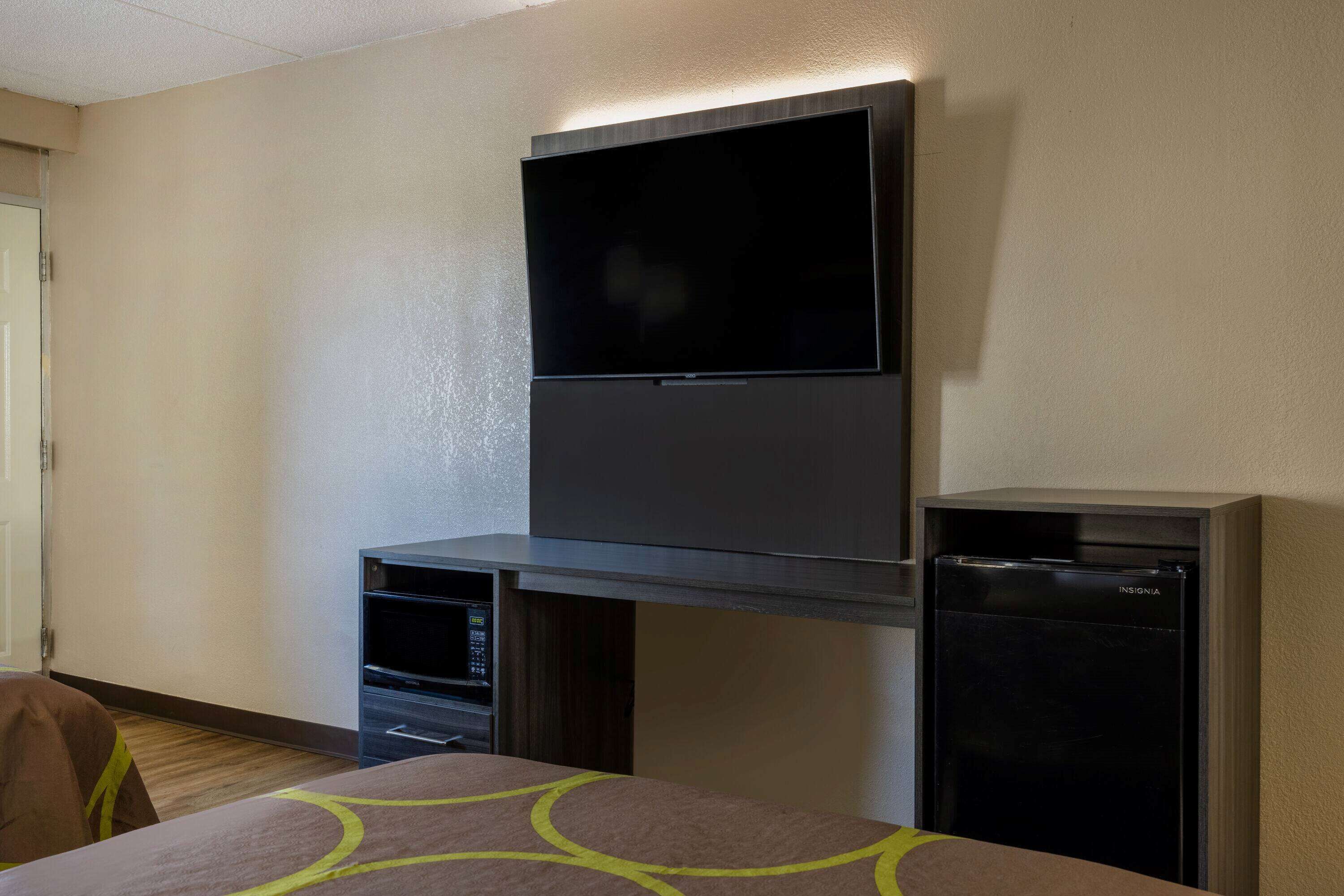 A hotel room with a flat-screen TV mounted on the wall, a small refrigerator, and a bed with a patterned bedspread.