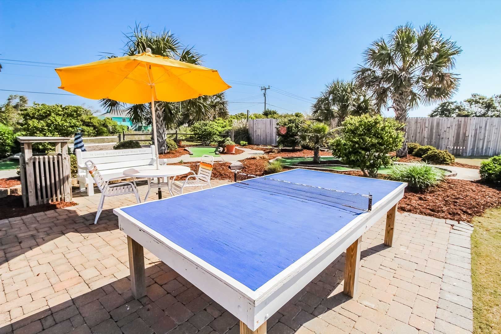 Un tavolo da ping pong all'aperto con superficie di gioco blu, circondato da un patio con pavimento in mattoni, un ombrellone giallo e diverse piante e alberi.