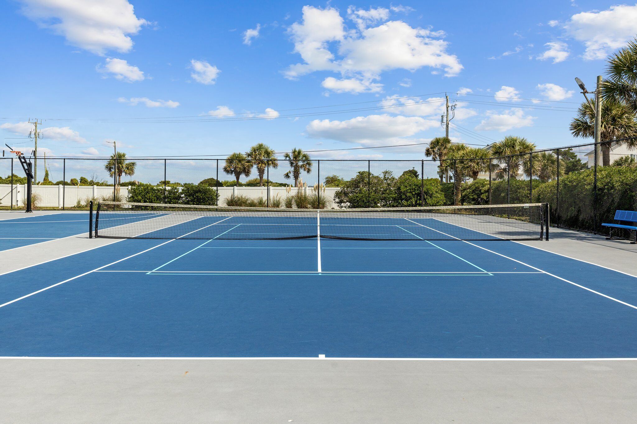 Een buitenterrein voor tennis met een blauwe ondergrond, omringd door een hek, palmbomen en een bank.