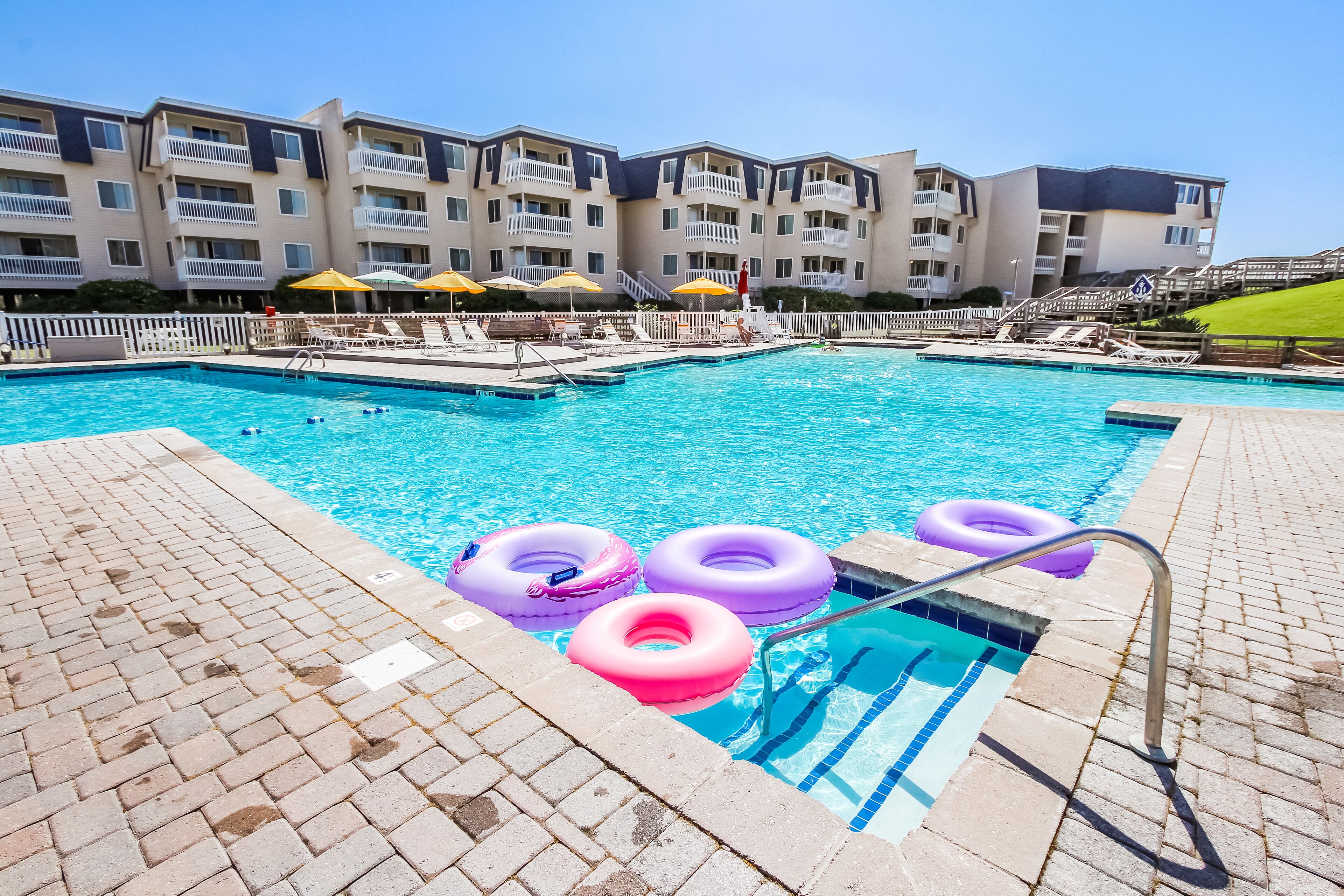 Une piscine entourée de chaises longues et de parasols, elle-même bordée d’un immeuble à plusieurs étages doté de balcons.