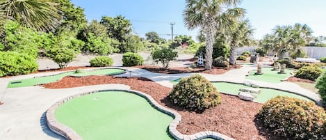 A miniature golf course with green putting greens, a brick border, and various shrubs and trees.
