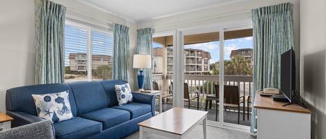 A modern hotel room with a blue sofa, a small table, and a balcony with a view of the city.